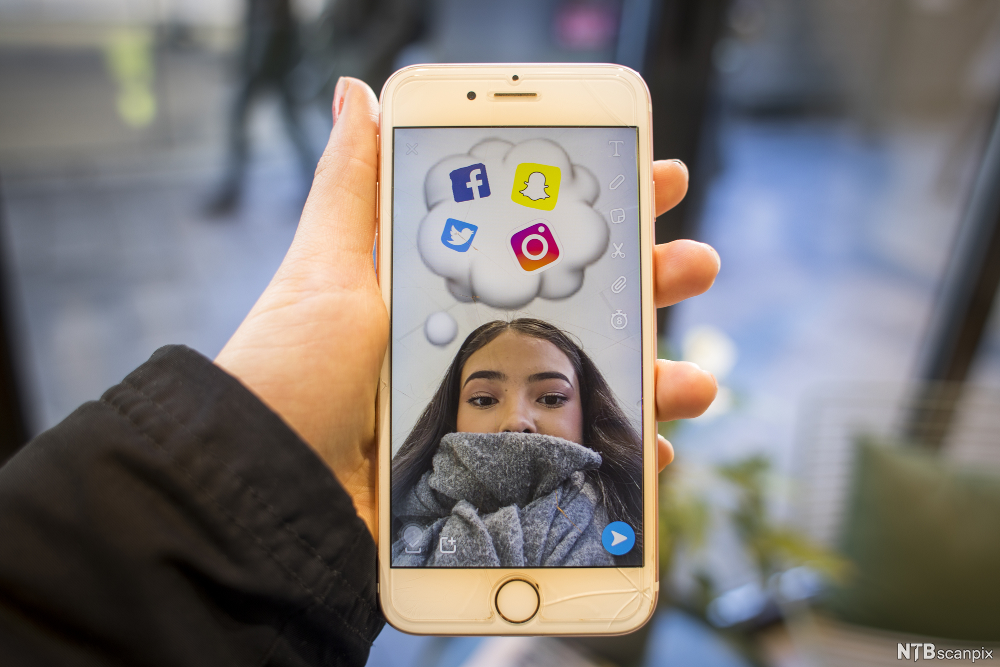 En hånd holder en mobiltelefon med bilde av ei jente som har logoer fra ulike sosiale medier i ei tenkeboble over hodet. Foto.