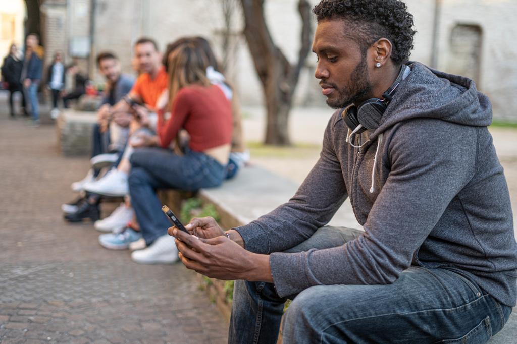 Ung mann sitter på en benk. Han er alene og ser på mobiltelefonen sin. I bakgrunnen sitter ei gruppe ungdommer som snakker sammen. Foto.