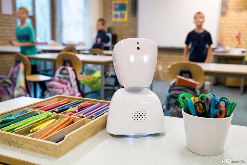 Robot forma som ein torso står på pult med fargeblyantar og sakser i eit klasserom. Elevar kan skimtast i bakgrunnen. Foto.