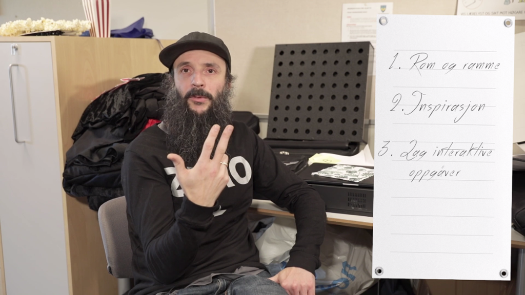 Mann med skjegg og kaps sitter ved en pult og holder opp tre fingre. På ei notisblokk til venstre i bildet står det "1. Rom og ramme", "2. Inspirasjon" og 3. Lag interaktive oppgaver". Skjermdump.