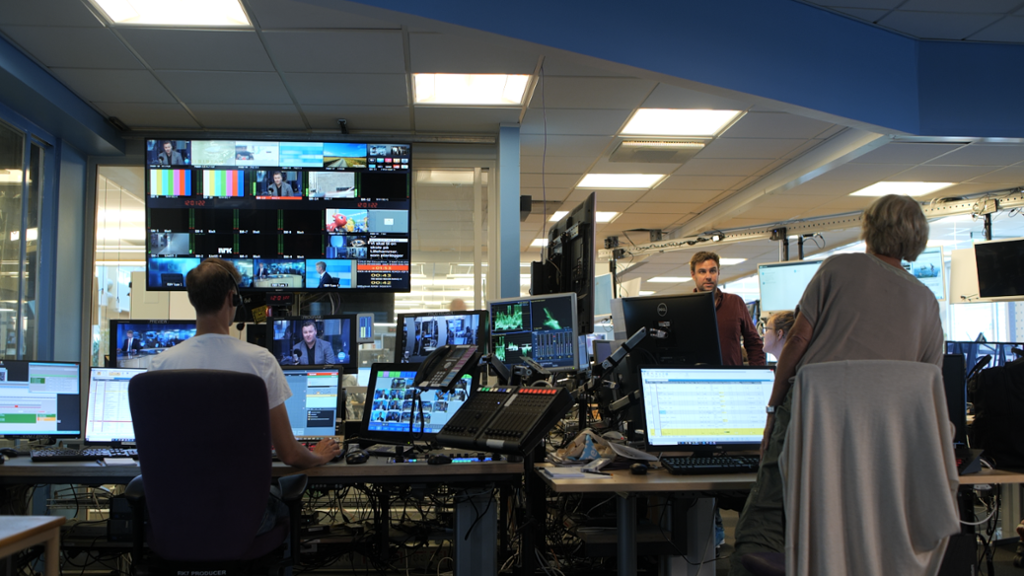 En moderne avisredaksjon med store skjermer og folk som sitter og jobber og kommuniserer. Foto.