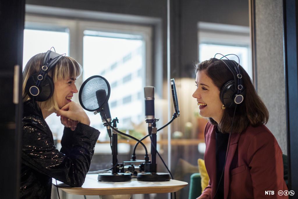 To kvinner sitter i et radiostudio. De har på seg hodetelefoner, og på bordet foran dem står det mikrofoner med popfilter. Foto.