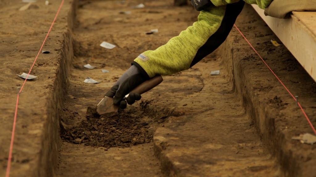 En person graver forsiktig i jorda med et lite verktøy som arkeologer bruker. Vedkommende ligger på magen, og vi ser bare hånden. Foto.