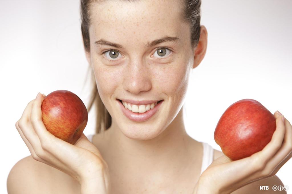  Ung, smilende kvinne med et rødt eple i hver hand. Foto.