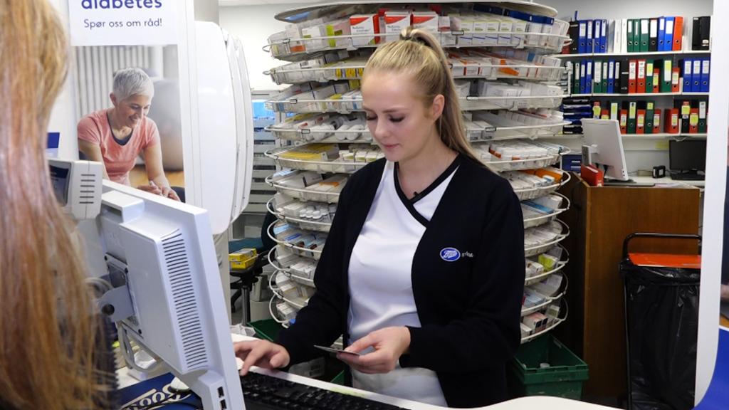 Apotektekniker står ved en pc og ekspiderer en kunde. Apotekteknikeren leser på id-kortet til kunden. Foto. 