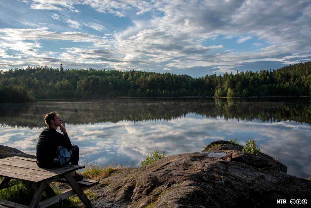 En mann sitter på et rastebord ute i naturen. Han ser ut over en innsjø. Foto.
