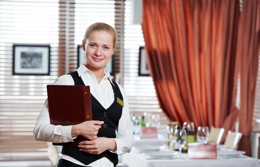 En kvinnelig servitør står foran et bord og holder en meny. Foto.