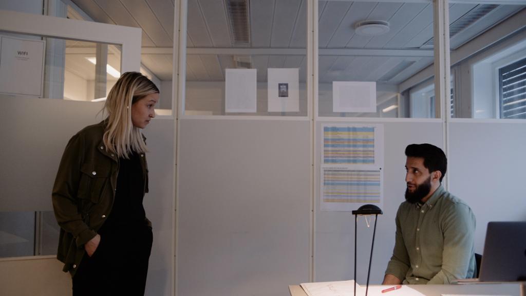 En kvinne står i en dør på et kontor og ser på en mann som sitter og forklarer noe. Foto.