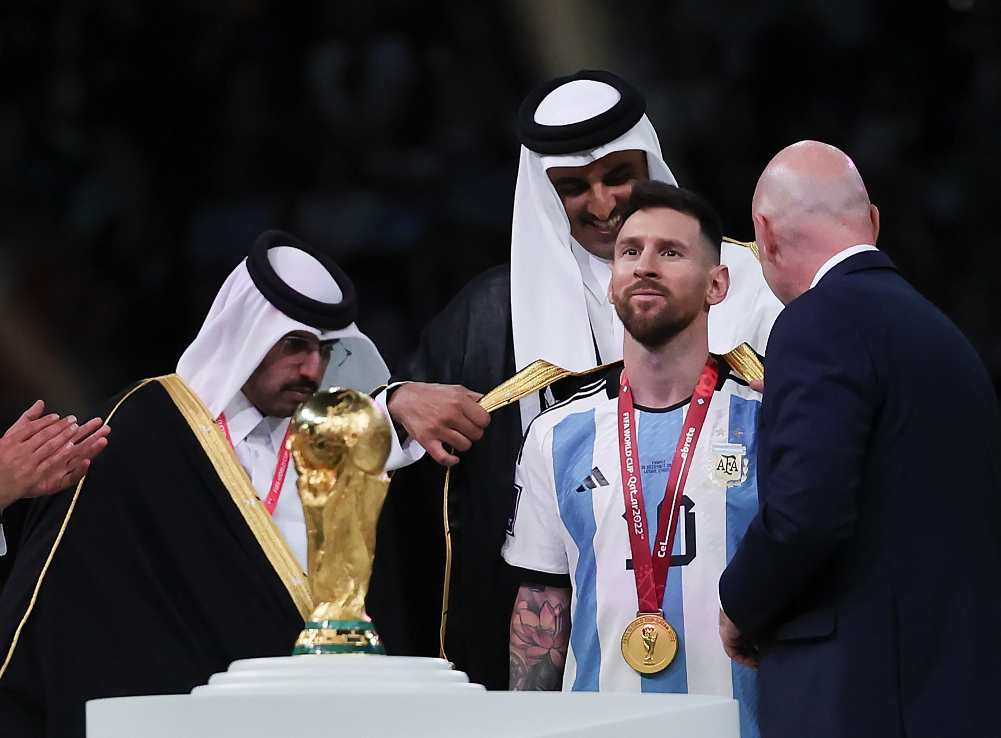 Ein mann i argentinsk fotballdrakt med gullmedalje på brystet får på seg ei kappe av to menn som er arabisk kledde og ein mann som er vestleg kledd. Foto.