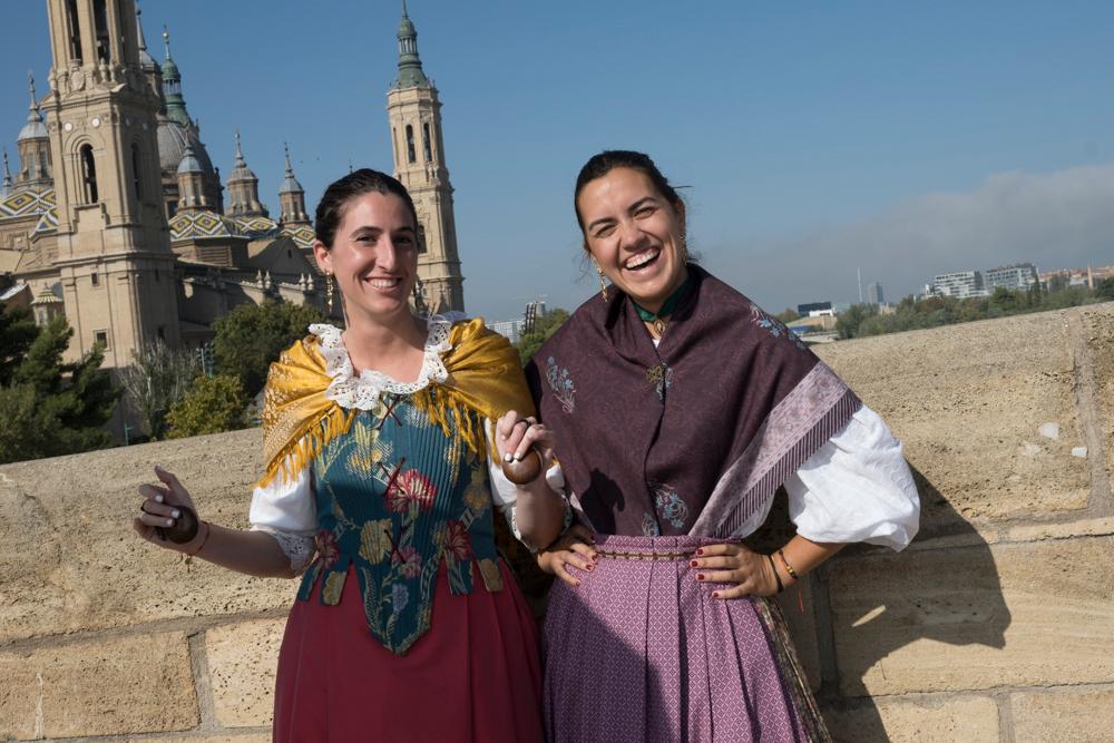 To glade kvinner i Zaragoza iført spansk folkedrakt. I bakgrunnen er kirken Basílica de Nuestra Señora del Pilar. Foto. 