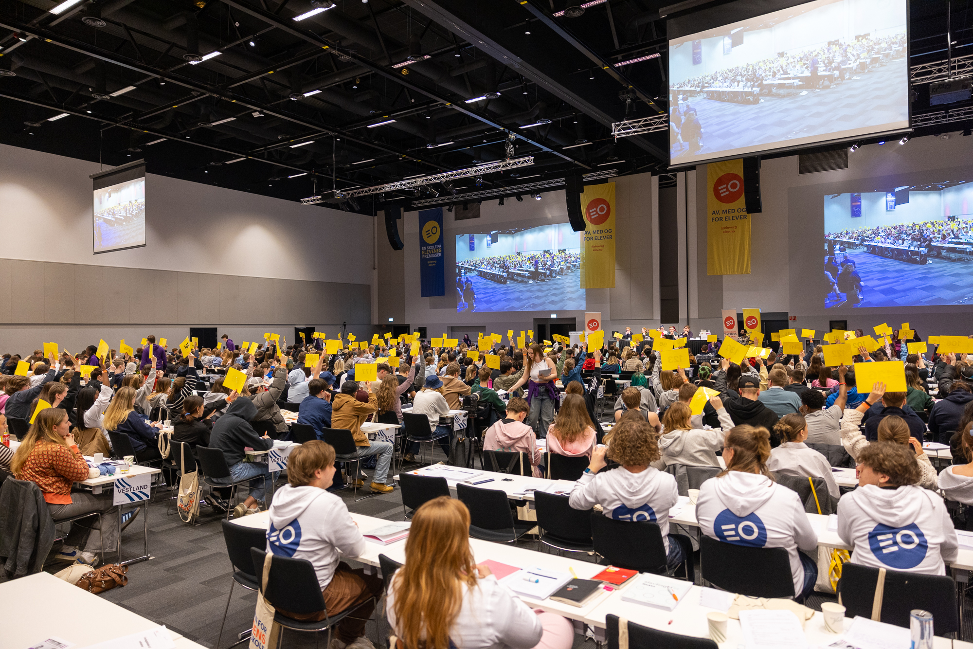 En stor forsamling ungdom sitter i en sal. Mange av dem holder opp et gult ark. Foto.