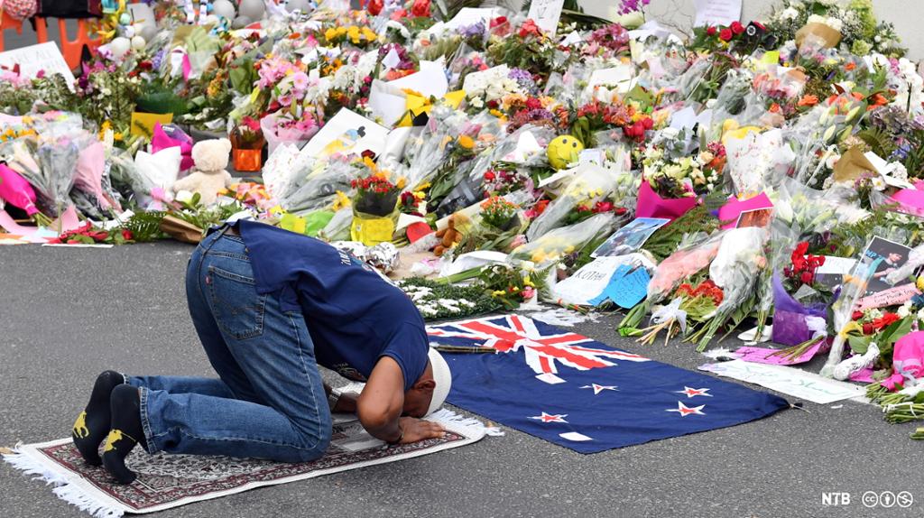 Photo: A Muslim worshipper prays at a makeshift memorial at the Al Noor Mosque in Christchurch, New Zealand. The memorial is packed with flowers, and a New Zealand flag is placed on the ground in front of the man.