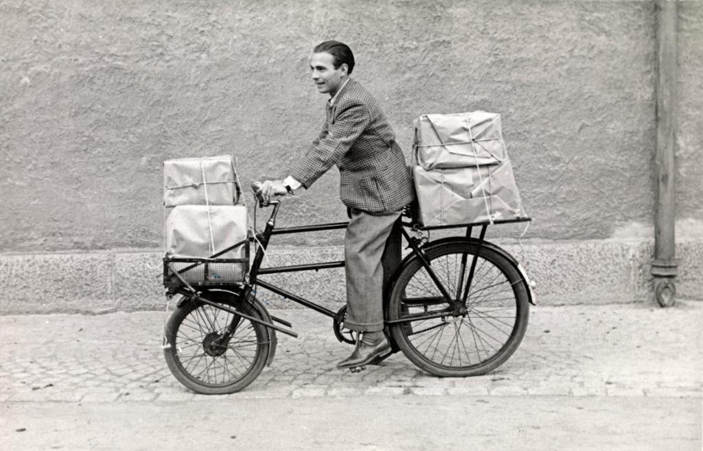 Ein mann på sykkel med fleire pakkar både framme og bak på sykkelen. Foto.