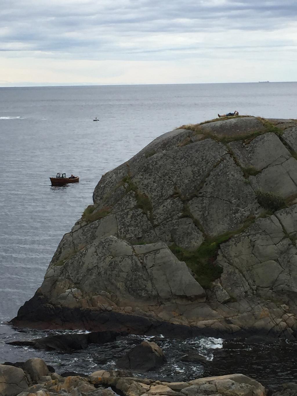 Båt på sjøen og mann som soler seg på svaberg. Foto.