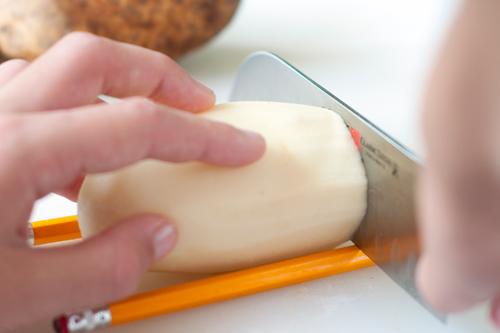 Nærbilde av kutting av potet. En skrelt potet ligger mellom to blyanter, slik at kniven stopper mot blyantene og ikke skjærer helt gjennom poteten. Foto.