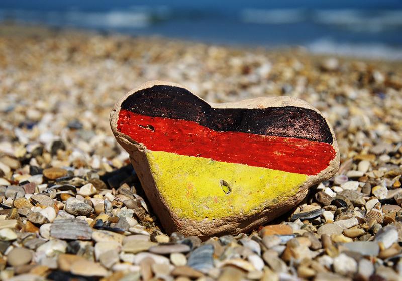 Foto som viser ei grusstrand i solskinn. I forgrunnen en hjerteforma stein der fargene til det tyske flagget er malt på.