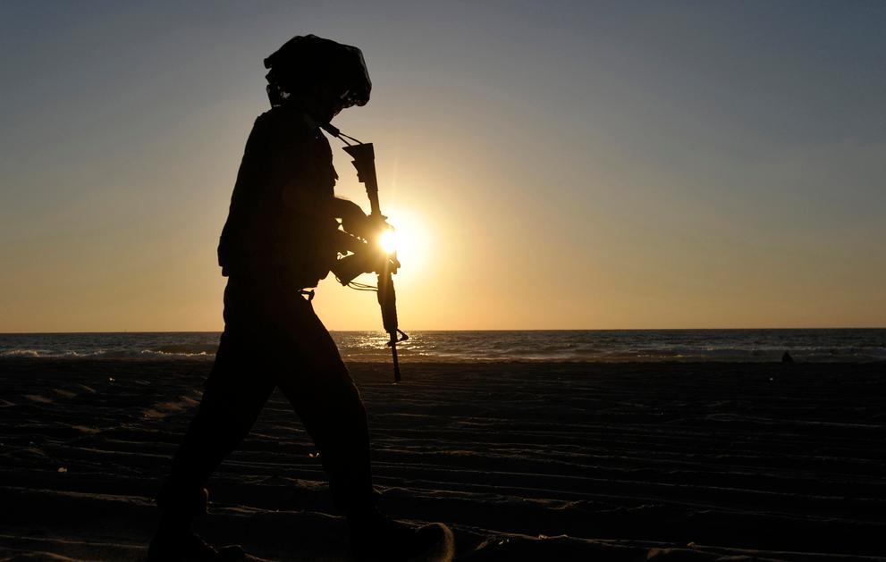 Mørk silhuett av ein gåande soldat med hjelm og gevær mot ein blågul, solfylt himmel ved havet. Foto.