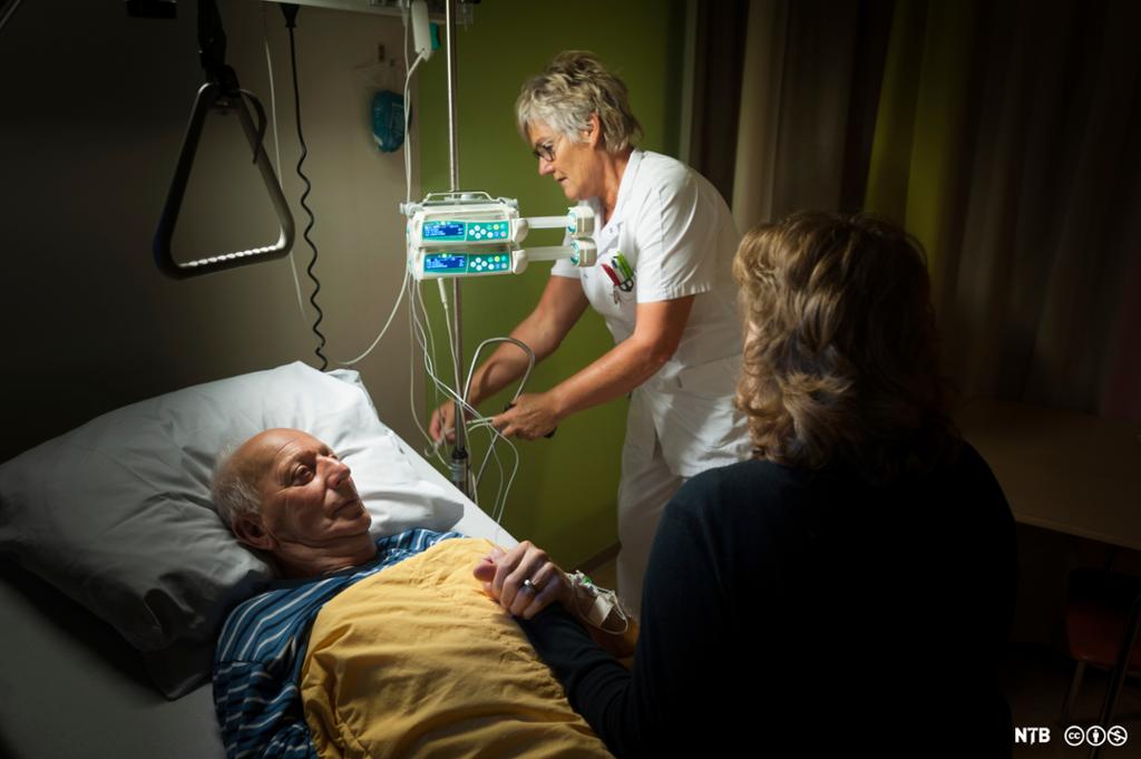 Sengeliggende pasient har besøk av pårørende som sitter på sengekanten hans mens en sykepleier ordner med infusjonspumpe. Foto. 