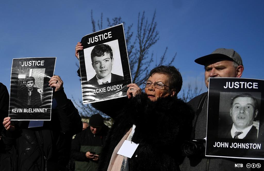 Photo: We see people holding up pictures of people with their names and the word 'justice'. There is a picture of a child with the name Kevil McElhinney, a teenager with the name Jackie Duddy, and a man with the name John Johnston. 