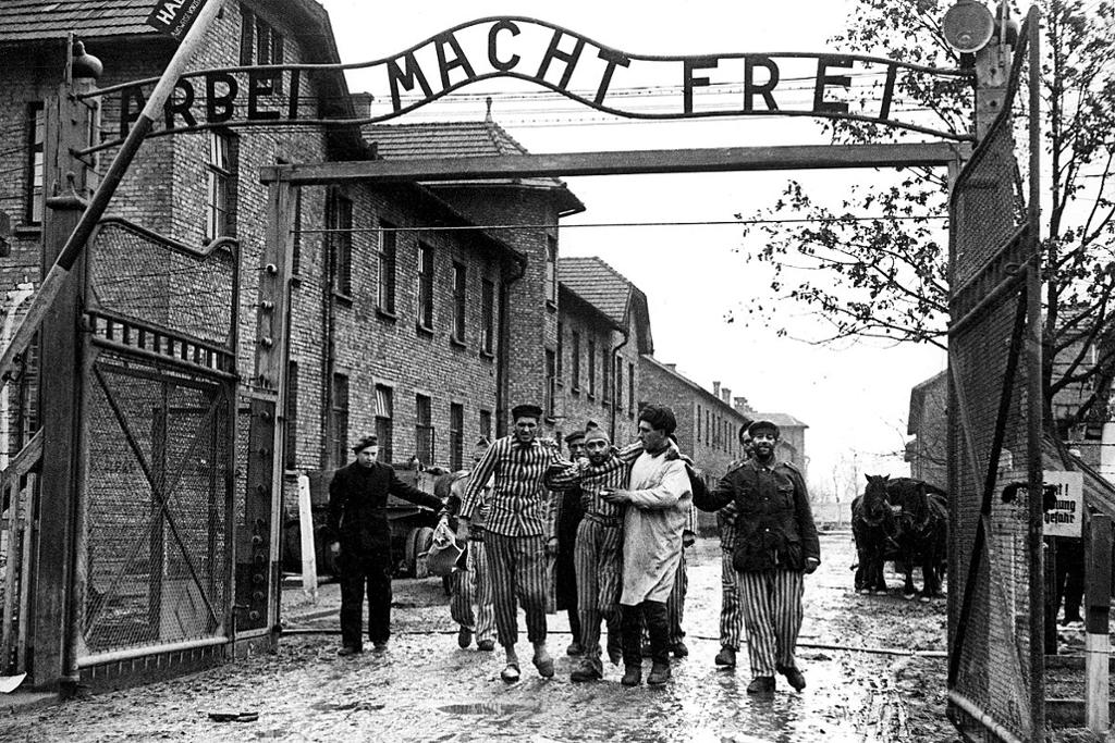 Gjenlevende fanger i stripete drakter ledes ut porten av sovjetiske soldater. Over porten står det på tysk: "Arbeit macht frei". Foto.