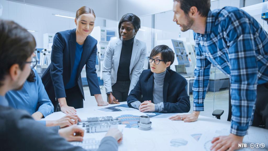 Photo: Adults standing around a table. Some are looking at objects on the table, some look at each other. They are of different ethnicities. 