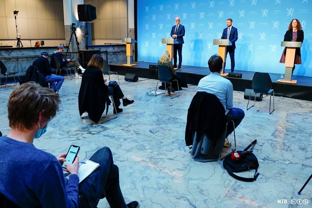 Pressekonferanse med tre personer på et podium. Journalister med munnbind sitter på stoler med stor avstand foran podiet. Foto.