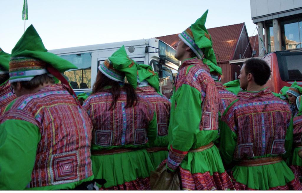 Ei gruppe voksne mennesker kledd i grønne etterligninger av samekofter står utenfor en buss. Foto.
