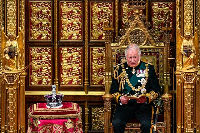 King Charles III delivers a speech in the House of Lords. He is sitting in a gilded chair with the crown sitting on a table next to him. The walls behind him are decorated in gold and red.