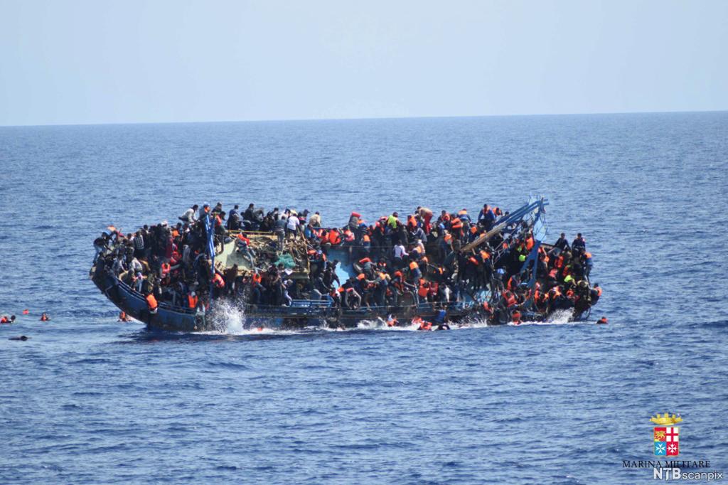 En overfylt båt kantrer. Om bord i båten er det svært mange mennesker, en del har redningsvester på og noen svømmer alt i sjøen. Foto.