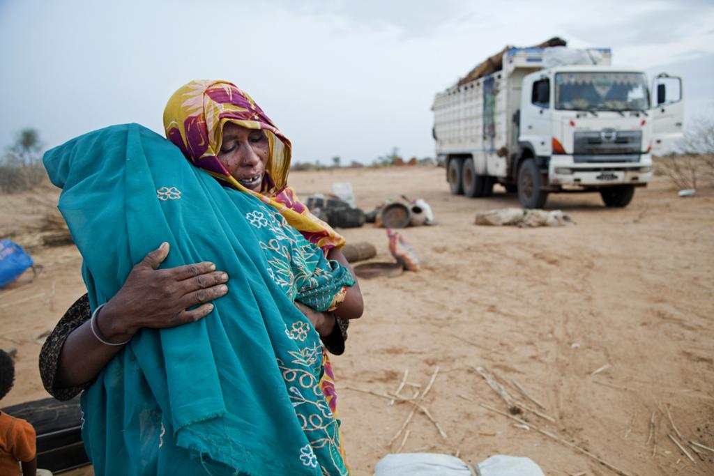 Photo: A woman wearing a yellow and purple headscarf is hugging a girl wearing a light blue headscarf. In the background there is a truck. They are in a sandy landscape. 