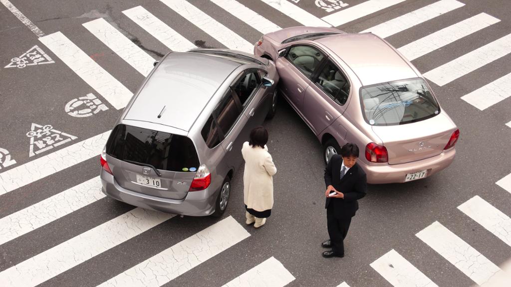 Sammenstøt mellom to biler i et veikryss. To personer står og ser på bilene som har kollidert. Foto.