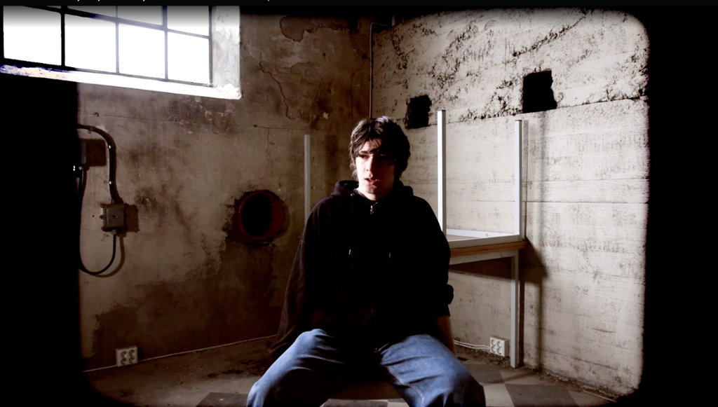 Gut som sit på ein stol i eit rom med betongveggar. Foto.
