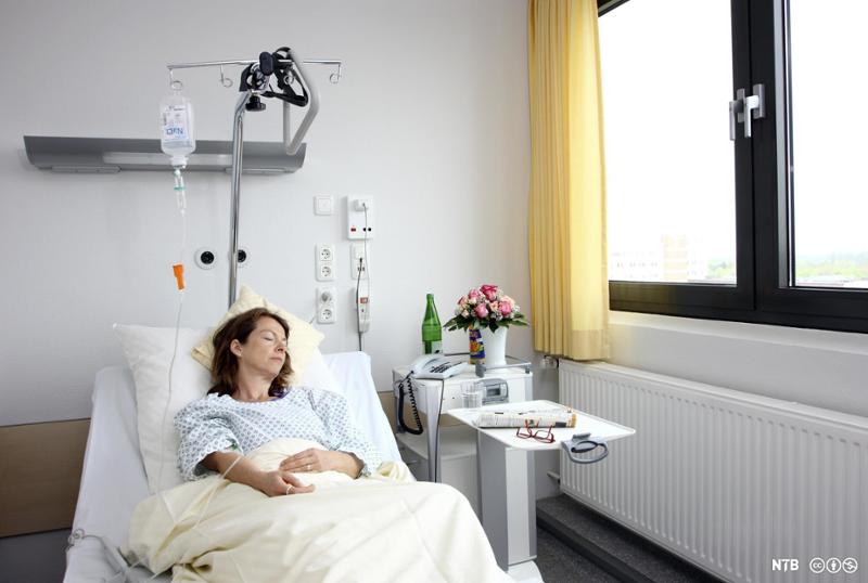 Kvinnelig pasient ligger i seng mens intravenøs behandling pågår. Foto.