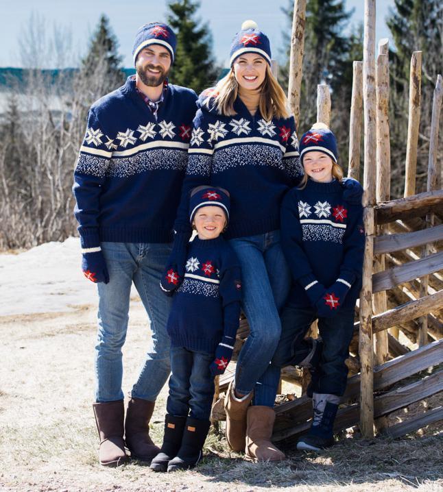 To voksne og to barn som har på seg mørkeblå strikkegensere og luer og votter i samme design. Alle smiler. De står ved en skigard, og det er trær og snø i bakgrunnen. Foto.