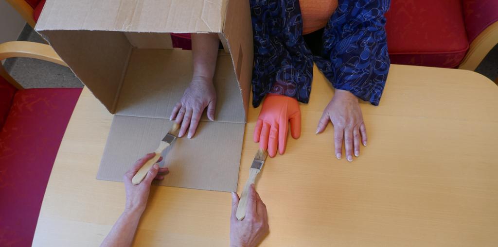 Forsøksoppsett der den ekte venstre hånda til forsøkspersonen er skjult og erstattet med ei synlig gummihånd. En annen person bruker pensler til å stryke fingrene på den ekte skjulte hånda og den falske hånda synkront. Illustrasjon.