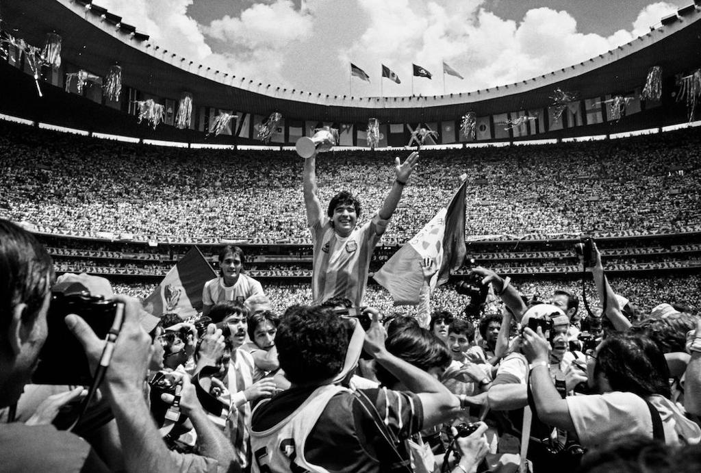 Fotballspilleren Diego Maradona løfter trofé omgitt av menneskemengde og tusenvis av tilskuere i bakgrunnen. Foto.