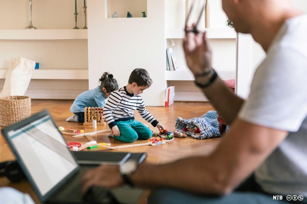 En mann med en bærbar PC sitter ved et bord og observerer barn som leker på gulvet. Foto.