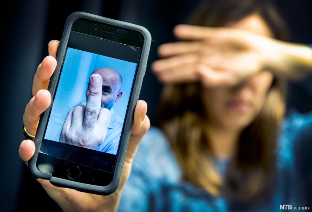 Opprørt kvinne viser fram mobil med bilde av mann som viser fingeren til kamera. Foto. 