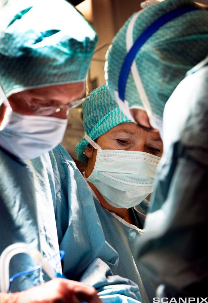 Nært bilde av et operasjonsteam. Tre leger er iført blå frakker, munnbind og hårnett. Den ene holder et operasjonsinstrument. Foto.