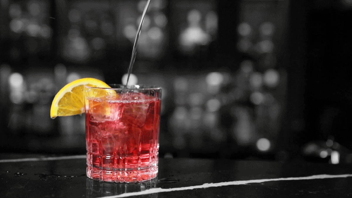 Raud drink i glas med appelsinbåt hengjande over glaskanten. Drinken står på ein bardisk. Foto.