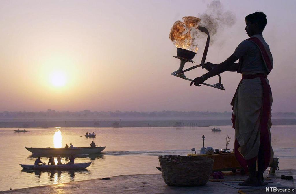 En mann står ved en elv og holder en skål med ild. På elven er flere mennesker ute i båt. Solen står lavt på himmelen. Foto.