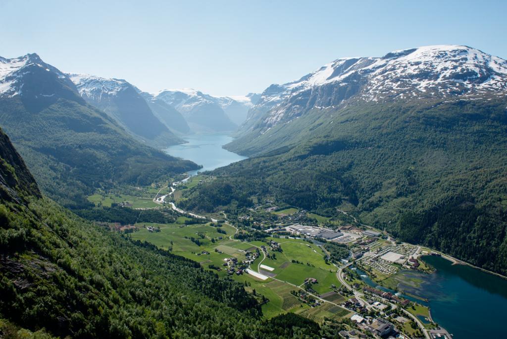 Bygd som ligger ved en fjord, med skogkledde åser på hver side. I bakgrunnen er det snødekte fjell på hver side av en smal innsjø. Foto.