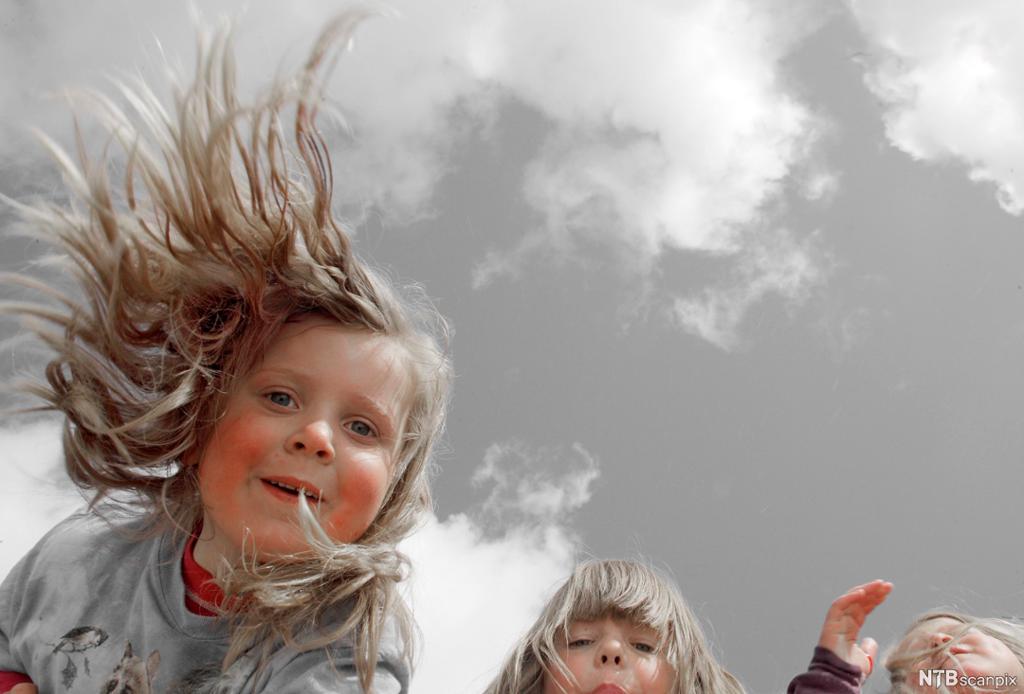 Bilde tatt nedenfra og opp som viser barn med vind i håret og himmelen med noen skyer. Foto.