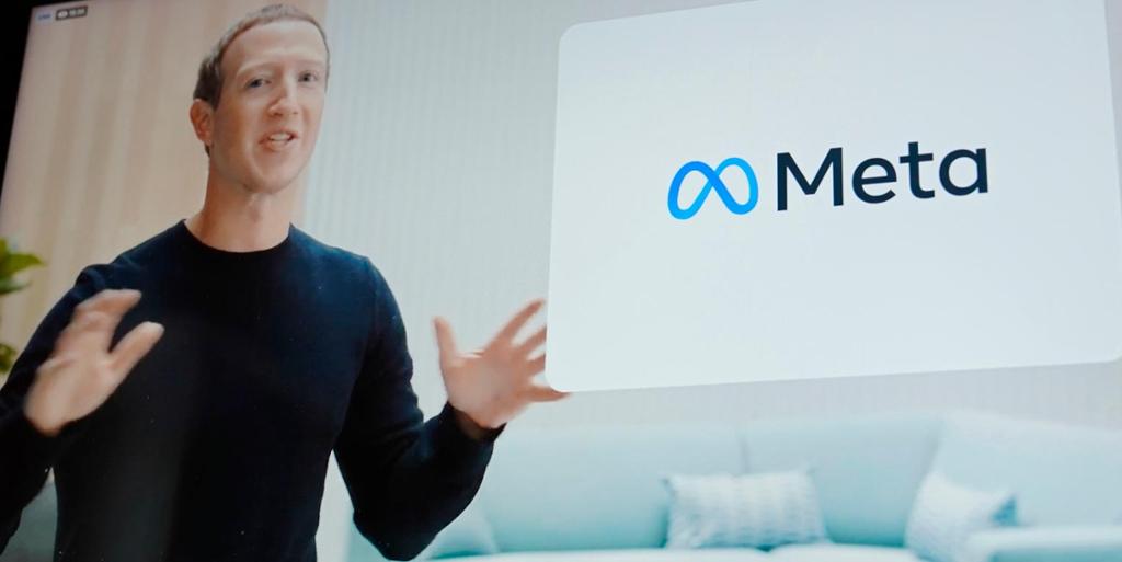 Foto av en skjerm som viser en mann i svart genser ved en digital logoplakat. Plakaten har hvit bakgrunn og teksten "Meta" i svart til høyre for en blå sløyfe som ligner et liggende åttetall eller det matematiske symbolet for en uendelig mengde.