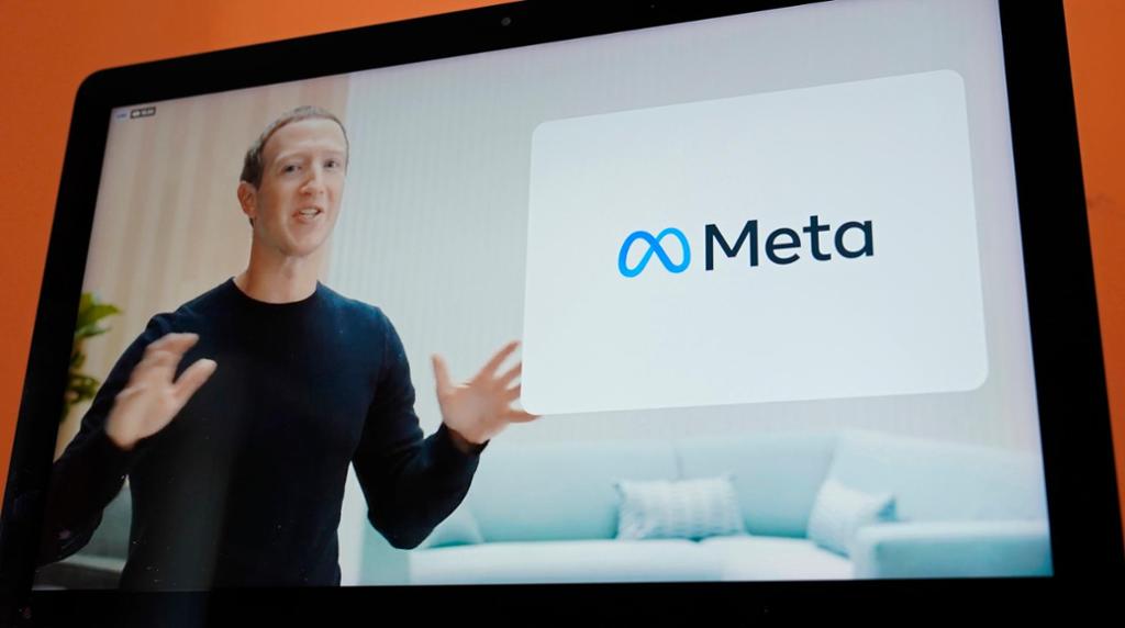 Foto av en skjerm som viser en mann i svart genser ved en digital plakat. Plakaten har hvit bakgrunn og teksten "Meta" i svart til høyre for en blå sløyfe som ligner et liggende åttetall eller det matematiske symbolet for en uendelig mengde.