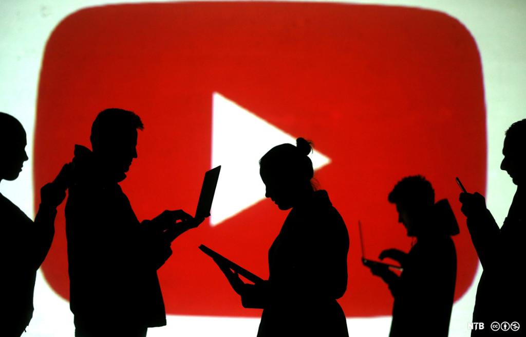 Foran YouTubes logo, en hvit play-knapp på rød bakgrunn, står silhuetter av fem personer med blikket festet i bærbare PC-er, nettbrett og mobiltelefoner. Illustrasjon.