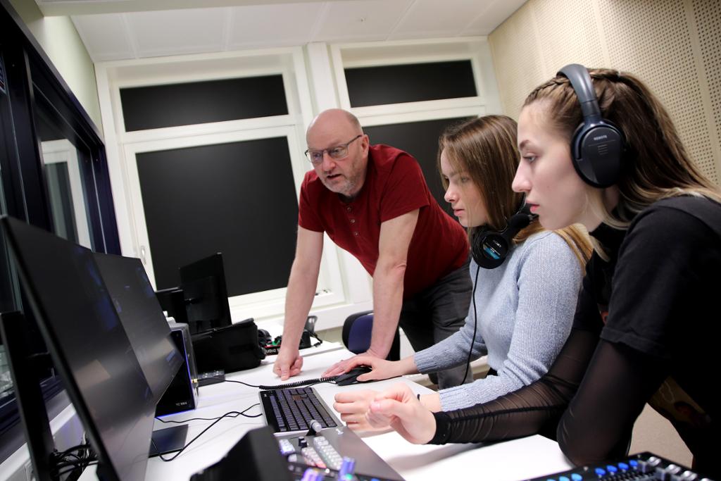Lærer og to elever foran skjermer, tastatur og bildemiks i et kontrollrom for tv-produksjon. Foto. 