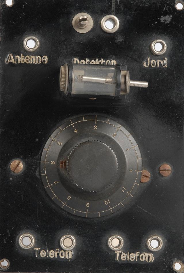 Fronten på et gammelt og litt rustent krystallapparat. Det er en svart boks med en dreieskive midt på og flere kontakter merket «Telefon», «Antenne», «Detektor» og «Jord». Foto.