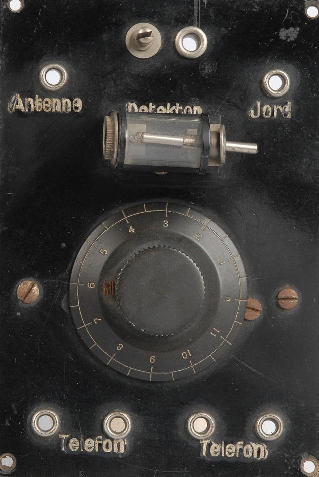 Fronten på eit gammalt og noko rustent krystallapparat. Det er ein svart boks med ei dreieskive midt på og fleire kontaktar merkte med «Antenne», «Detektor», «Jord» og «Telefon». Foto.