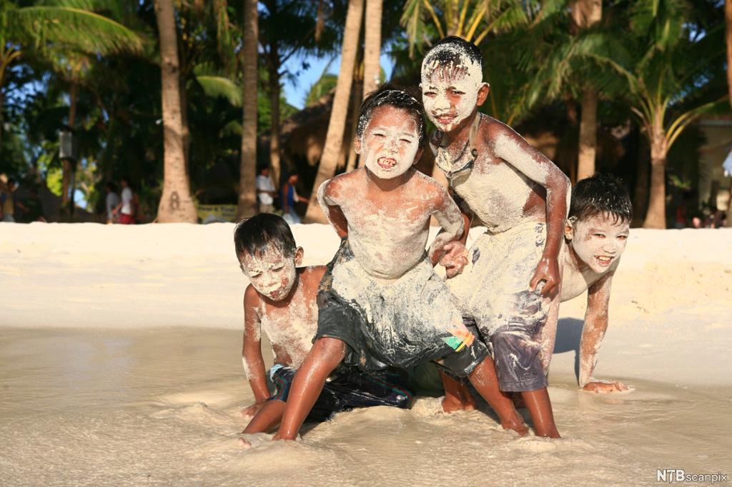 Fire gutter som leker i vannkanten på ei kjent strand. De er våte og har rulla seg i sanda, så de er dekket av sand. Foto.